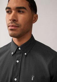 Camicia nera con bottoni in tessuto testurizzato, caratterizzata da un colletto a punta e bottoni bianchi; include un piccolo logo ricamato sul petto.
