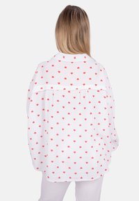 White shirt with red heart patterns, oversized design, lightweight fabric, visible collar, drops at the back, textured surface.