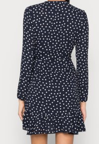 Frau, die ein marineblaues Kleid mit langen Ärmeln, weißen Punkten und einem gerafften Saum trägt, von hinten gezeigt.