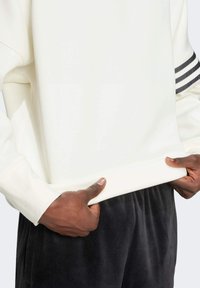 Sweatshirt blanc à manches longues et poignets côtelés, avec trois rayures foncées sur le haut de la manche et un ourlet lisse sans embellissement.