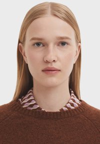 Brauner Wollpullover mit rundem Halsausschnitt, über einem gemusterten Hemd mit lila, weißen und braunen geometrischen Formen getragen. Glatte Textur.