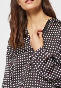 Kvinna som bär en svart blus med vit och rosa rutmönster, håller vid halsringningen nära svarta knappar, brunt hår som faller över axlarna.