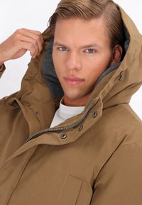 Brun parka-jakke med en tekstureret overflade, stor hætte, grå foring, frontlynlås og trykknapper. Modellen justerer hætten.