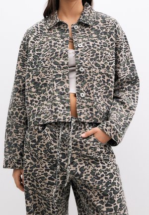 Femme portant une veste et un pantalon assortis à imprimé léopard avec un top court blanc, main dans la poche, sur fond blanc.