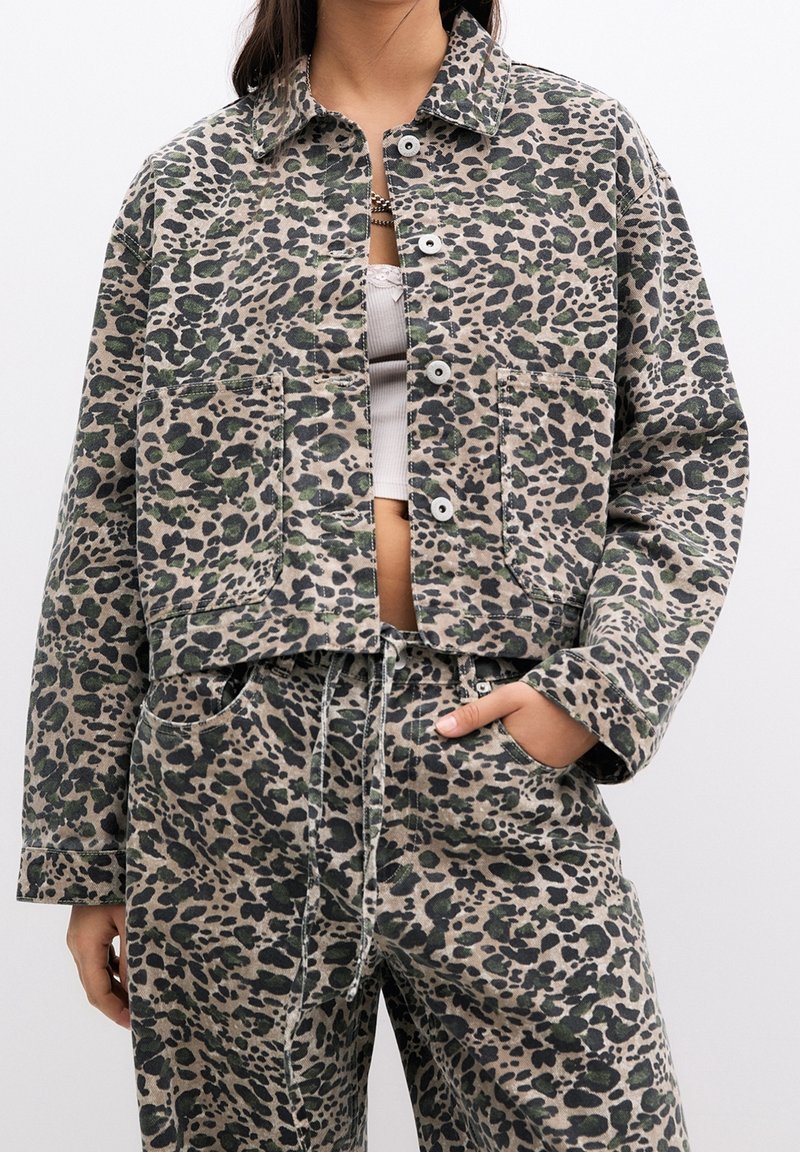 Femme portant une veste et un pantalon assortis à imprimé léopard avec un top court blanc, main dans la poche, sur fond blanc.