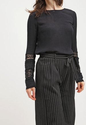 Femme portant un chemisier noir à manches en dentelle et un pantalon noir à rayures taille haute, noué avec une ceinture en tissu.