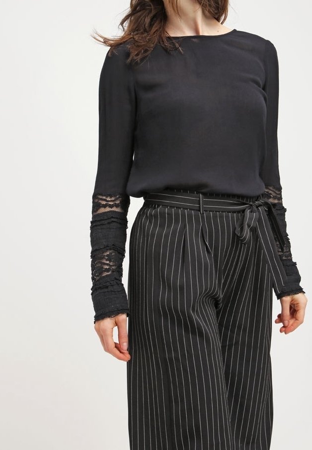 Femme portant un chemisier noir à manches en dentelle et un pantalon noir à rayures taille haute, noué avec une ceinture en tissu.