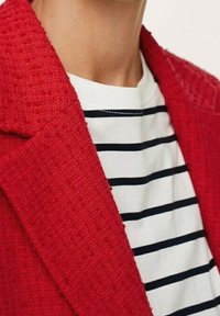Veste rouge texturée avec un col cranté portée sur un haut à col rond blanc rayé de bleu marine. Accent sur la matière et les détails du design.
