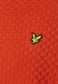 Tessuto arancione testurizzato con un motivo a griglia, caratterizzato da un logo di uccello ricamato in nero e giallo in basso a sinistra.