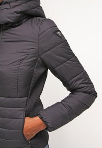 Svart vadderad jacka med dragkedja framtill, huva och texturerade sidopaneler. Har en diamantlogotyp på axeln och ribbade ärmslut.