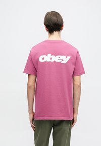 Růžové bavlněné tričko s velkým bílým logem "obey" tištěným na zadní straně. Materiál má jemnou strukturu a volný střih.