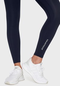 Marina leggings med en slät yta, prydda med vit text "SQUATPROOF". Matchas med vita träningsskor med nätdesign och snörning.