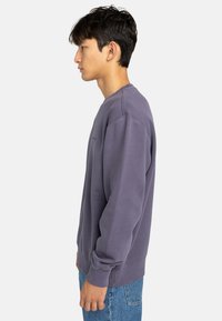 Purple fleece crewneck sweatshirt with ribbed cuffs and hem, featuring a relaxed fit, shown on a model in profile. Blue denim worn below.