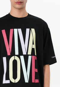Μαύρη μπλούζα με κοντό μανίκι, που διαθέτει έντονα, πολύχρωμα γράμματα "VIVA LOVE" με υφή. Κοντά μανίκια, στρογγυλή λαιμόκοψη, λογότυπο στον μανίκι.
