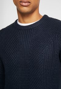 Marinblå stickad tröja med ett strukturerat mönster och ribbad rundad halsringning. Tyget har en mjuk, något glänsande yta.