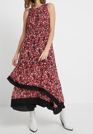Robe midi rouge fleurie sans manches avec bordure noire portée avec des bottines blanches sur une femme debout sur un fond uni.