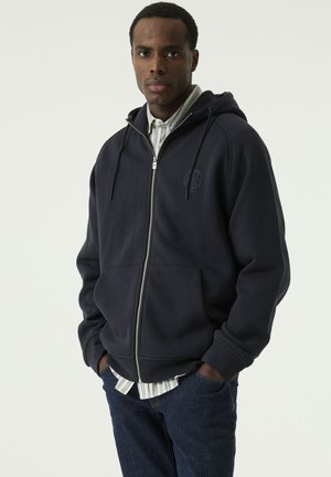 Man staat recht vooruit, draagt een donkerblauwe hoodie met rits over een overhemd met kraag en een blauwe jeans, handen in de zakken, neutrale achtergrond.