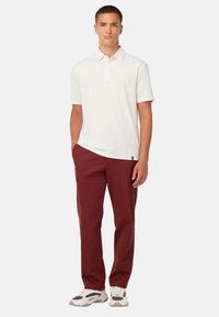 Bílá polo košile s límcem a třemi knoflíčky, v kombinaci s vínovými kalhotami. Model stojí v neformálních sportovních botách.