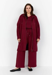 Burgunderfarbene Lounge-Wear mit einer Reißverschlussjacke mit Taschen, passenden Jogginghosen mit Kordelzug und schwarzen Slippers. Weicher, entspannter Schnitt.