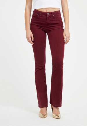 Kvinde iført bourgognefarvede, højtaljede, tætsiddende jeans med gyldne spidse hæle og en hvid crop top mod en ensfarvet hvid baggrund.