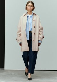 Heller rosa übergroßer Trenchcoat mit braunen Knöpfen, getragen über einem blau gestreiften Hemd und dunklen Denim-Hosen, kombiniert mit schwarzen spitzen Schuhen.