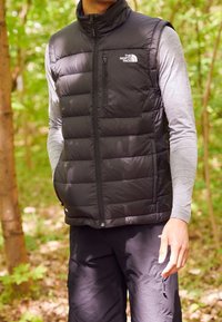 Muž v černé vyteplené vestě s logem The North Face, šedém tričku s dlouhým rukávem a černých kalhotách v lesním prostředí.