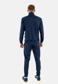 Marineblaue Trainingsanzug aus glattem Stoff, bestehend aus einer zipfbaren Jacke und enganliegenden Hosen. Schwarze Seitenstreifen verleihen den Beinen Details.