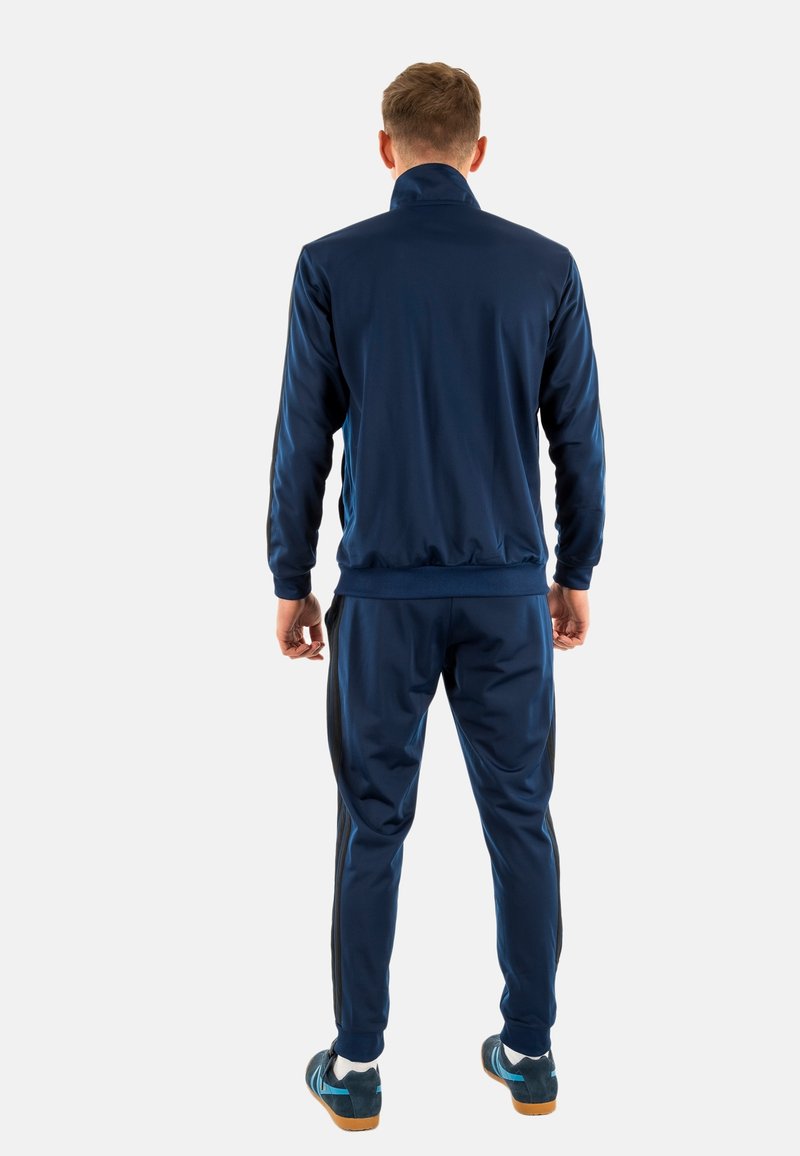 Survêtement bleu marine en tissu lisse, composé d'une veste zippée et d'un pantalon ajusté. Des bandes noires sur les côtés apportent une touche de détail le long des jambes.
