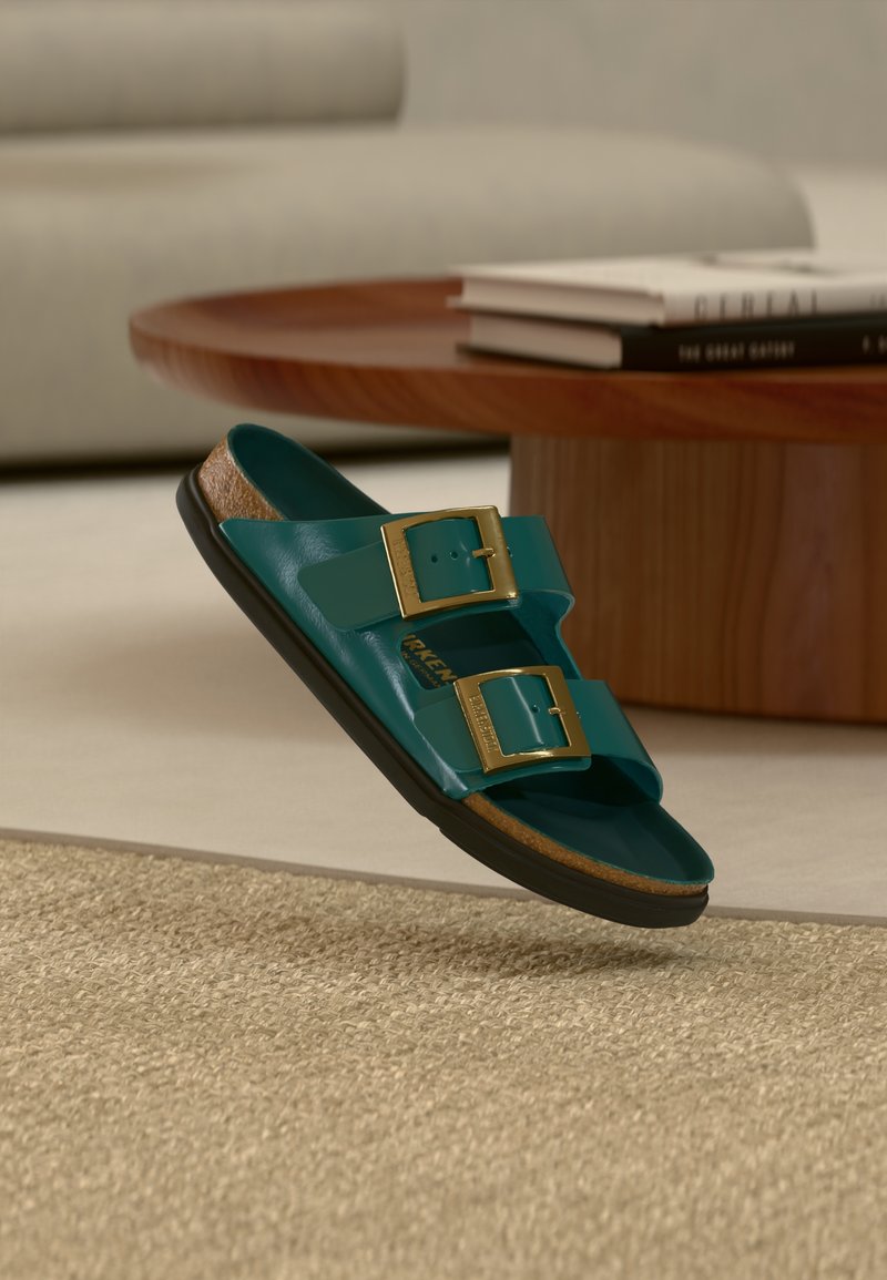 Τυρκουάζ σανδάλι τύπου slip-on με δύο χρυσά αγκράφια να αιωρείται πάνω από μπεζ χαλί, ξύλινο στρογγυλό τραπεζάκι σαλονι ού με στοιβαγμένα βιβλία και φόντο γκρι καναπέ.