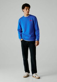 Άνδρας που στέκεται με τα χέρια στις τσέπες, φορώντας ένα φωτεινό μπλε φούτερ, μαύρο παντελόνι και λευκά αθλητικά παπούτσια σε απλό φόντο.