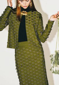 Costume vert olive texturé avec des motifs en V saillants de couleur violette, comprenant une veste cintrée et une jupe assortie. Accents d'accessoires dorés inclus.