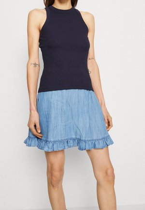 Frau trägt ein ärmelloses, marineblaues, geripptes Oberteil und einen hellblauen Denim-Minirock mit gerafftem Saum, sie steht vor einem schlichten hellen Hintergrund.