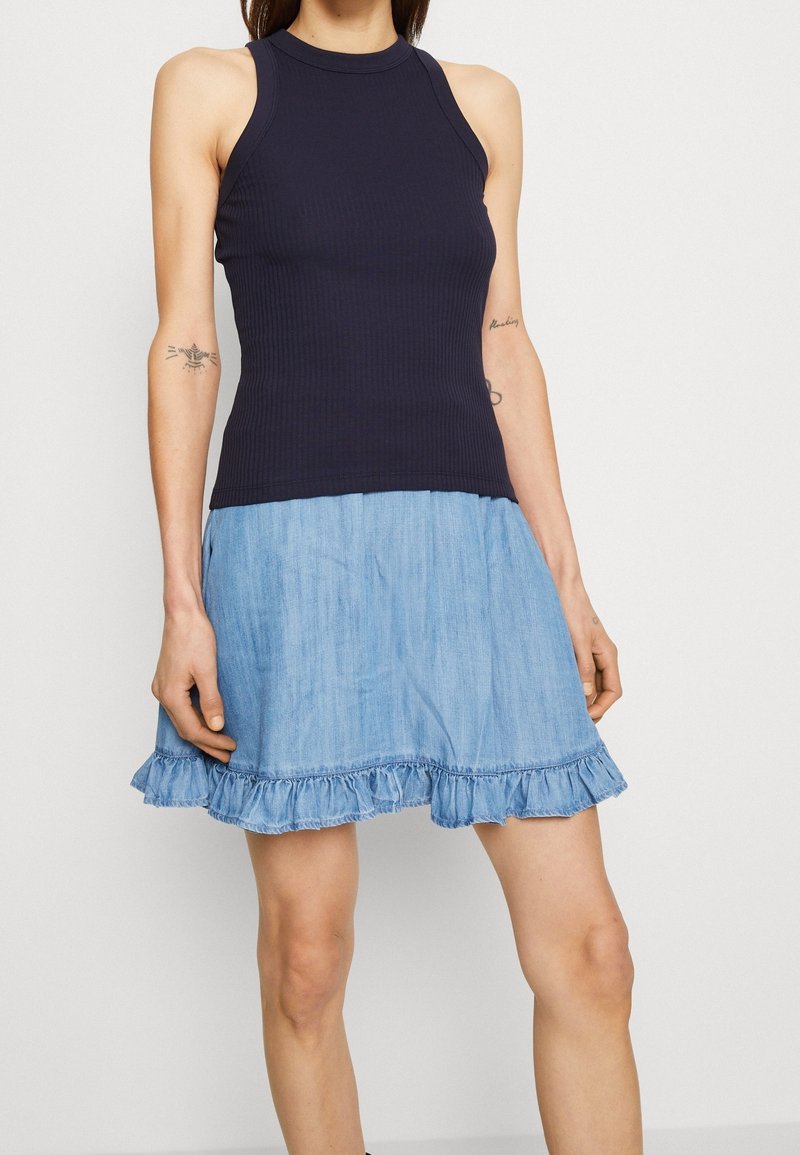 Femme portant un haut sans manches bleu marine côtelé et une mini-jupe en denim bleu clair avec un ourlet à volants, debout devant un fond clair uni.
