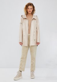 Beige, waterproof jacket with a hood, front zipper, two patch pockets, and silver accents. Paired with light beige trousers and sneakers.