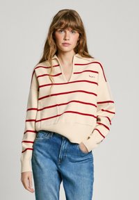 Maglione crema con righe rosse orizzontali, colletto e tasca frontale; indossato con jeans blu. Texture morbida, design casual.
