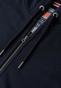 Mørkeblå zip-up hoodie lavet af glat bomuld med kontrasterende hvide og røde striber indvendigt, sølv lynlås og broderet logo.
