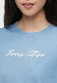 Gaiši zila sporta krekla ar apaļu kakla izgriezumu un baltu izšūtu "Tommy Hilfiger" logo uz krūtīm, kas izgatavots no mīksta materiāla.