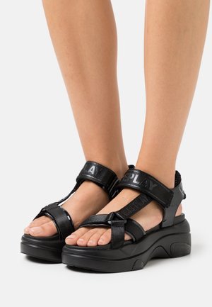 Sandales à plateforme noires avec une semelle en caoutchouc, dotées de sangles ajustables, d'accents brillants et d'un dessus en tissu texturé.