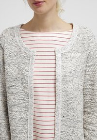 Cardigan texturé gris clair avec encolure ronde, devant ouvert et bordure blanche contrastante ; porté sur un haut à rayures rouges.