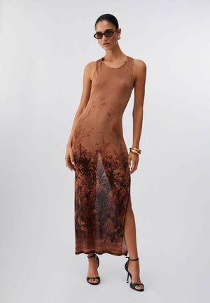 Femme portant une robe longue marron sans manches avec un motif floral noir, des sandales à talons hauts noires, des lunettes de soleil et des bracelets dorés sur un fond uni.