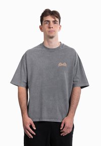 Camiseta cinza oversize com decote redondo e mangas curtas, com um desenho gráfico castanho claro no peito, feita de tecido macio.