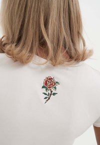 Hvid bomulds T-shirt med en rød og pink broderet rose med grønne blade, placeret på ryggen nær halsudskæringen.