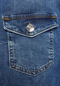 Napattu sinisen denim-taskun nappilukko, jossa on keltaiset ompeleet farkkukankaassa, näyttäen tekstuuria ja kankaan kudontatietoja.