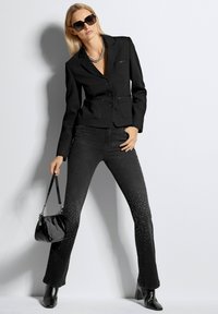 Schwarzer strukturierter Blazer mit zwei Fronttaschen, kombiniert mit flared schwarzen Jeans, die mit Strasssteinen verziert sind, und einer glänzenden schwarzen Handtasche.