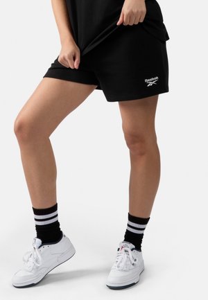 Personne portant un short noir Reebok, des chaussettes noires à rayures blanches et des baskets blanches Reebok, debout avec une jambe légèrement fléchie.