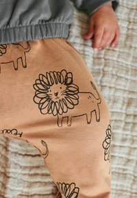 Lysbrune bukser med sorte løveillustrationer og et tekstureret gråtaljebånd. Blødt stof synligt mod en mønstret overflade.