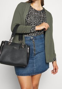 Nainen, jolla on päällään vihreä bleiseri, musta-valkoinen kuvioitu toppi, edestä vetoketjulla varustettu farkkuhame, ja hän kantaa mustaa Guess-käsilaukkua olkapäällään.