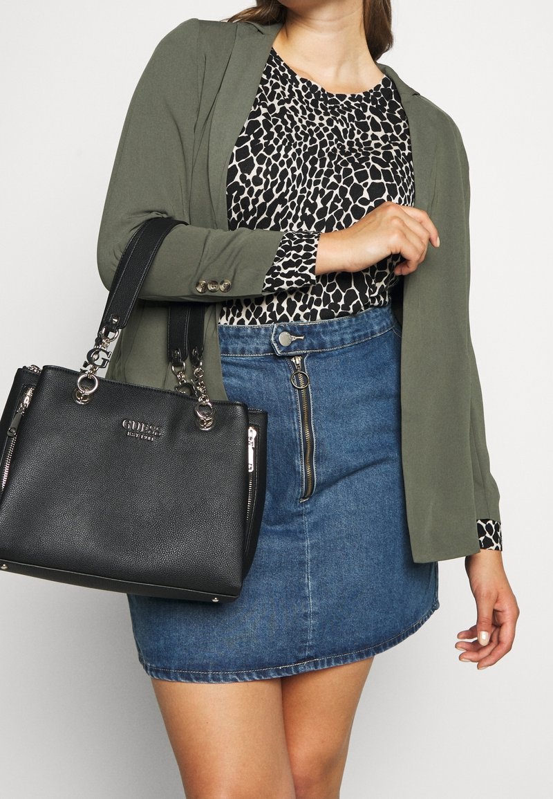 Nainen, jolla on päällään vihreä bleiseri, musta-valkoinen kuvioitu toppi, edestä vetoketjulla varustettu farkkuhame, ja hän kantaa mustaa Guess-käsilaukkua olkapäällään.