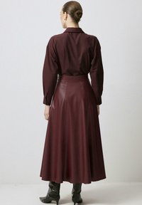 Sieviete ar matiem sasuktos bunā, ģērbusies garā bordo ādas svārkā, atbilstošā blūzē un melnās papēžkurpēs, stāv pretī baltai sienai.