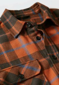 Chemise en flanelle boutonnée avec un motif à carreaux orange, vert foncé et bleu, avec un col et une poche munie d’un bouton noir.